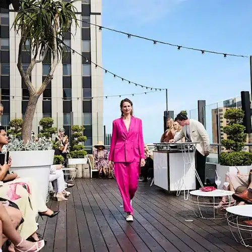 A woman in a bright pink suit confidently walks along a wooden deck, showcasing her stylish outfit.