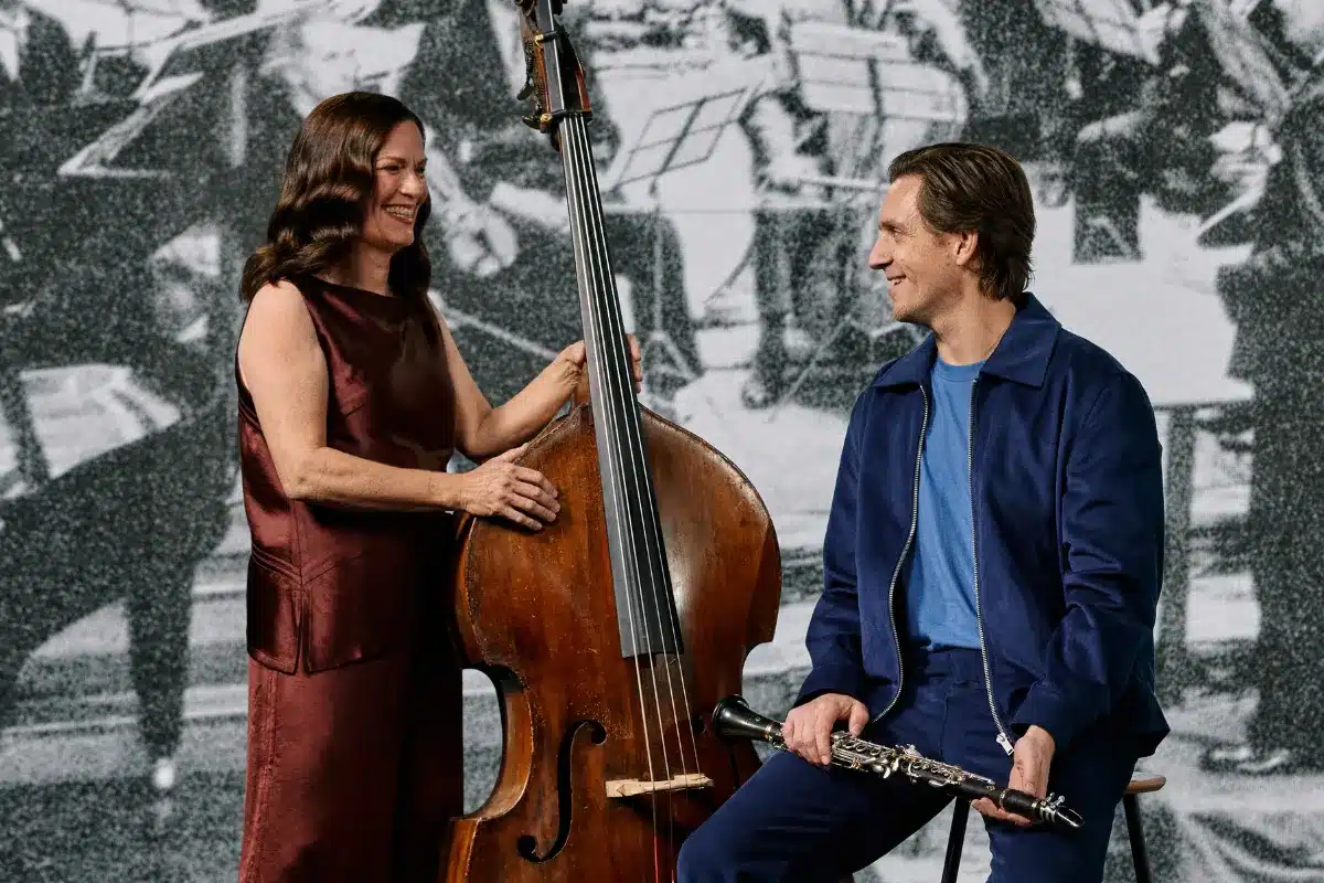 A man and woman sit side by side, playing a double bass together in a cozy setting.