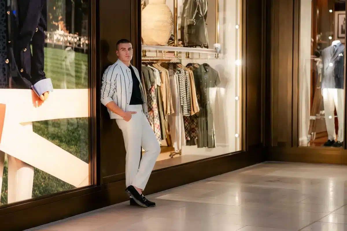 A man stands in front of a store window showcasing various clothing items on display.