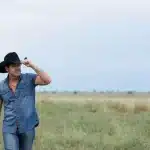 A man wearing a cowboy hat stands confidently in a wide, open field under a clear blue sky.