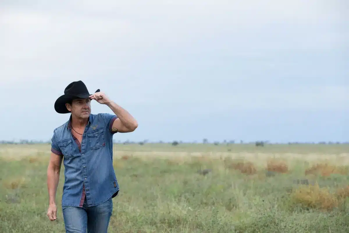 A man wearing a cowboy hat stands confidently in a wide, open field under a clear blue sky.