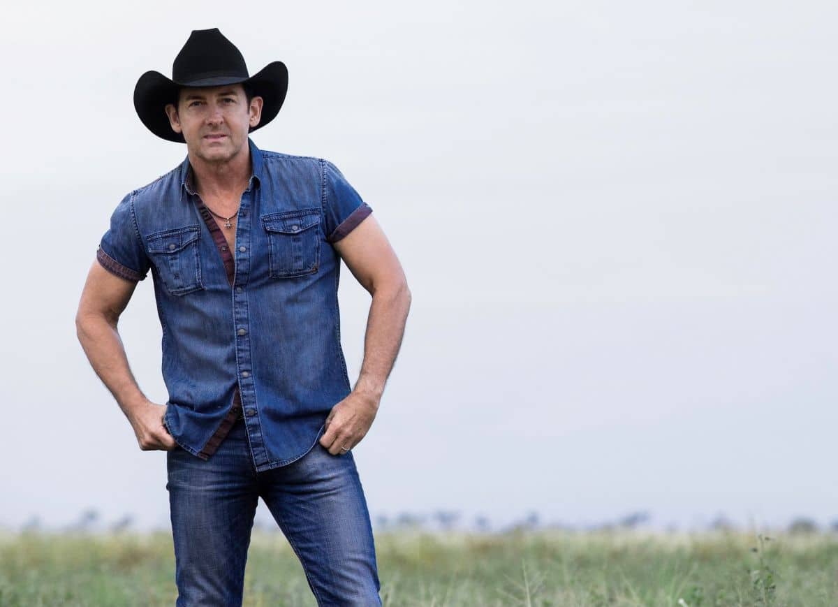 A man wearing a cowboy hat stands confidently in a wide, open field under a clear blue sky.