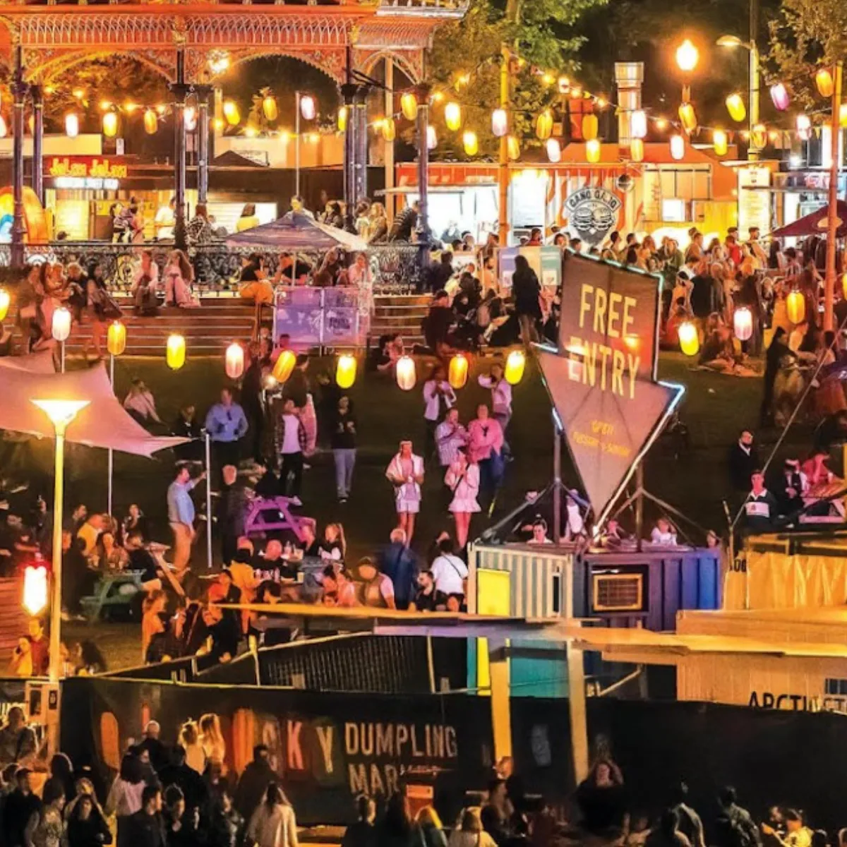 A lively crowd of people enjoying an outdoor fair at night, illuminated by colorful lights and festive decorations.