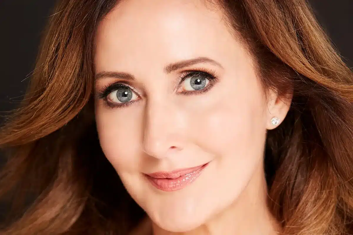 A woman with long brown hair and blue eyes gazes thoughtfully into the distance.