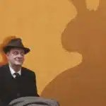 A man in a hat and suit sits on a chair, casting a shadow of a rabbit on the ground beside him.
