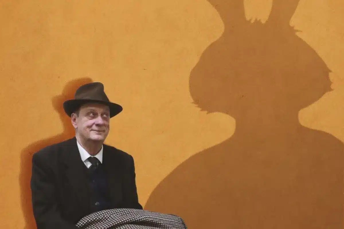 A man in a hat and suit sits on a chair, casting a shadow of a rabbit on the ground beside him.