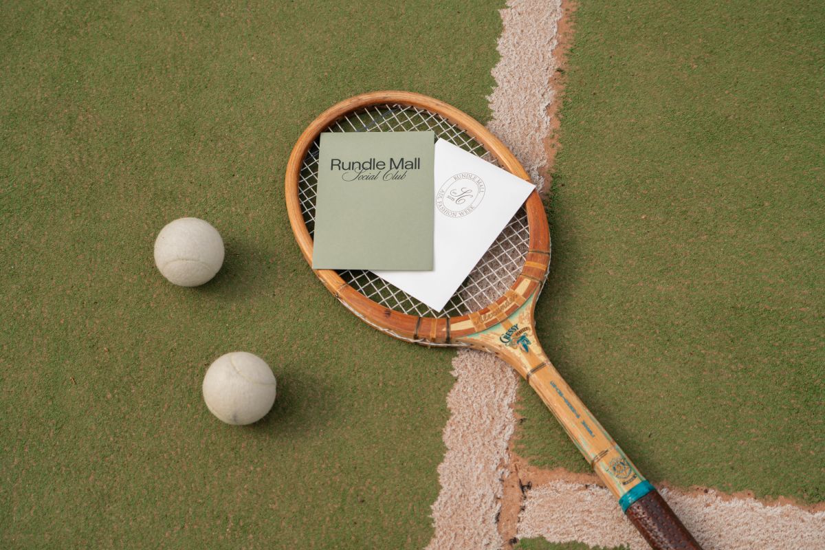 A tennis racket and several balls are placed on a vibrant green tennis court.
