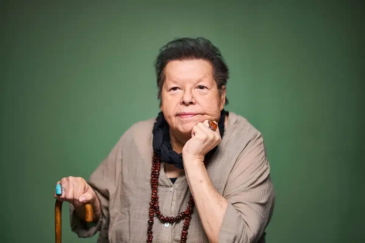 An older woman with gray hair, using a cane, wearing a colorful necklace.