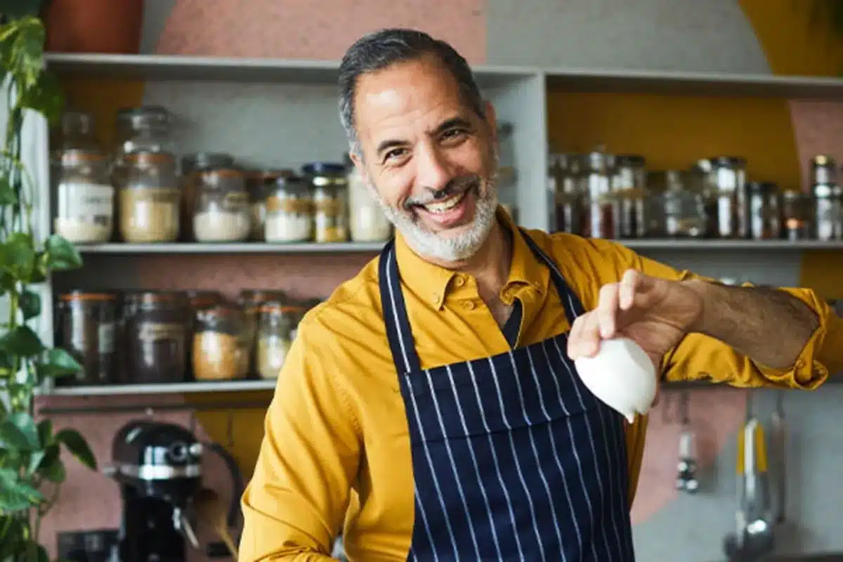 A man wearing an apron smiles as he prepares to cook in a kitchen.