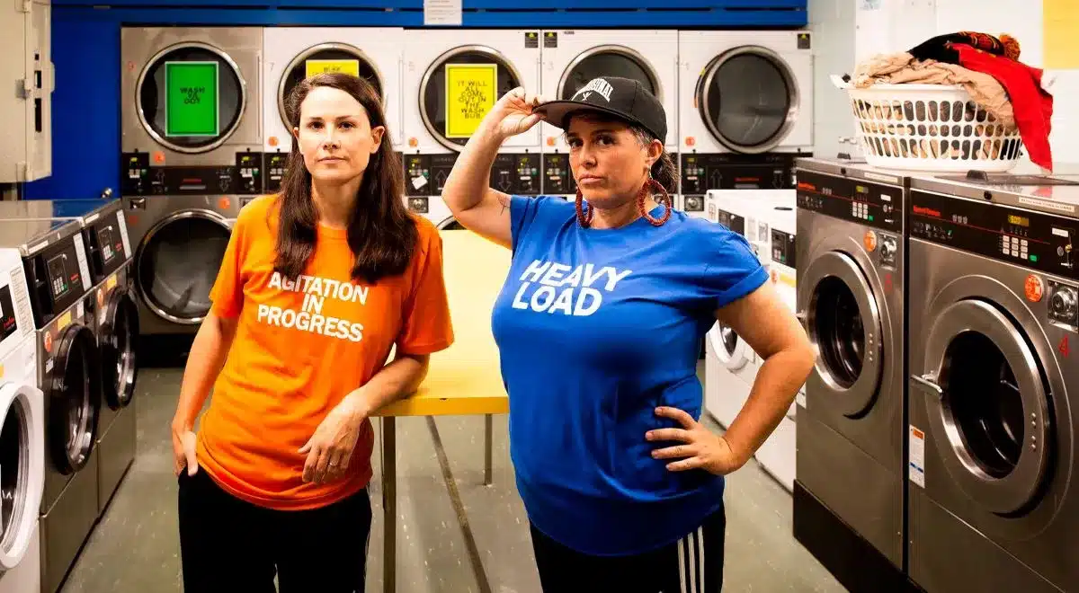 Two women stand in a laundry room, one loading a washing machine while the other observes.