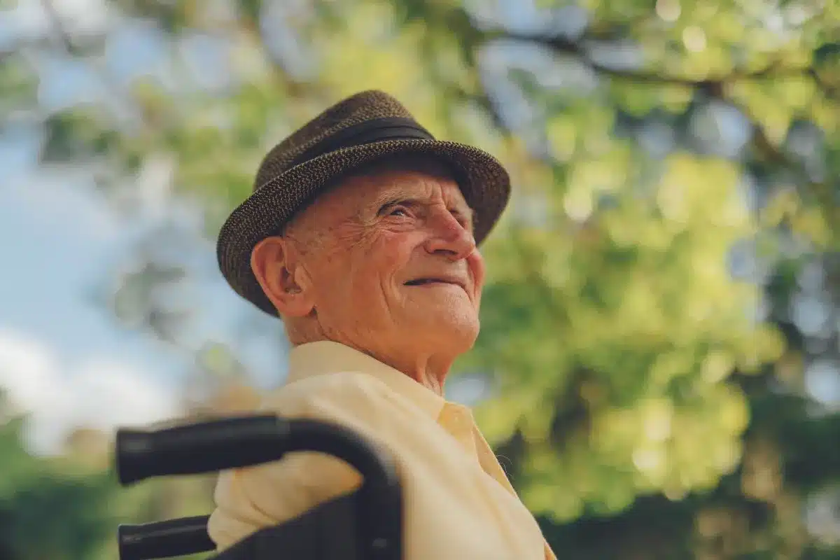 An older man wearing a hat sits in a wheelchair, looking content and relaxed.