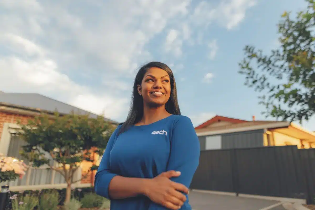 A woman in a blue shirt stands in front of a house, smiling and looking towards the camera.