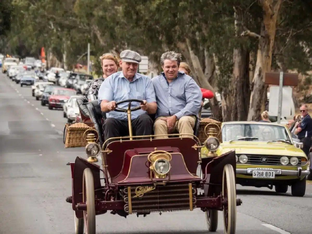 Three people, two men and a woman, are in an antique car traveling along a picturesque road.