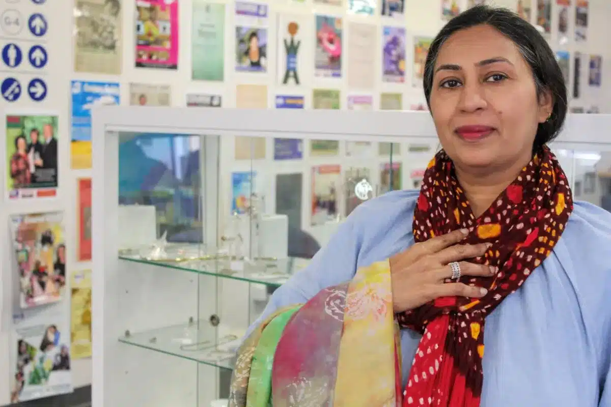 A woman in a blue shirt and red scarf stands in front of a vibrant display of colorful scarves.