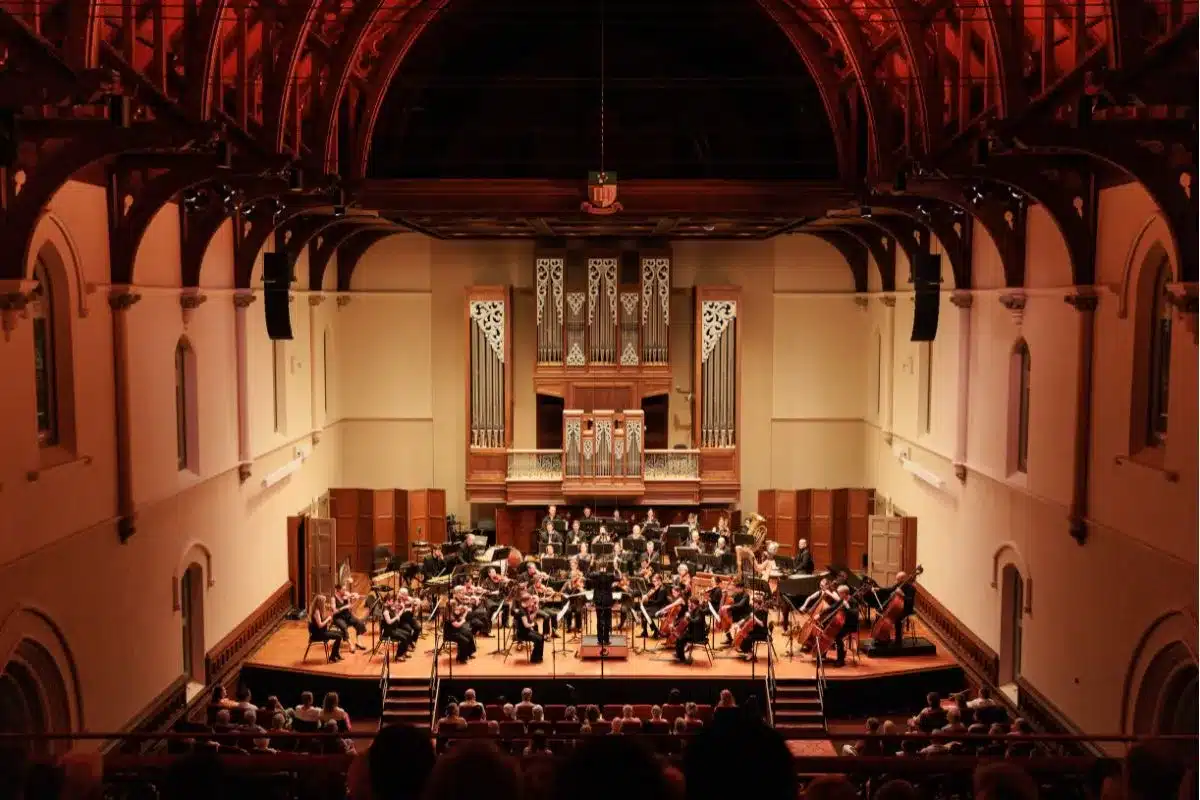 A large orchestra is seen performing in a music hall, with a magnificent organ adding to the solemn ambiance.