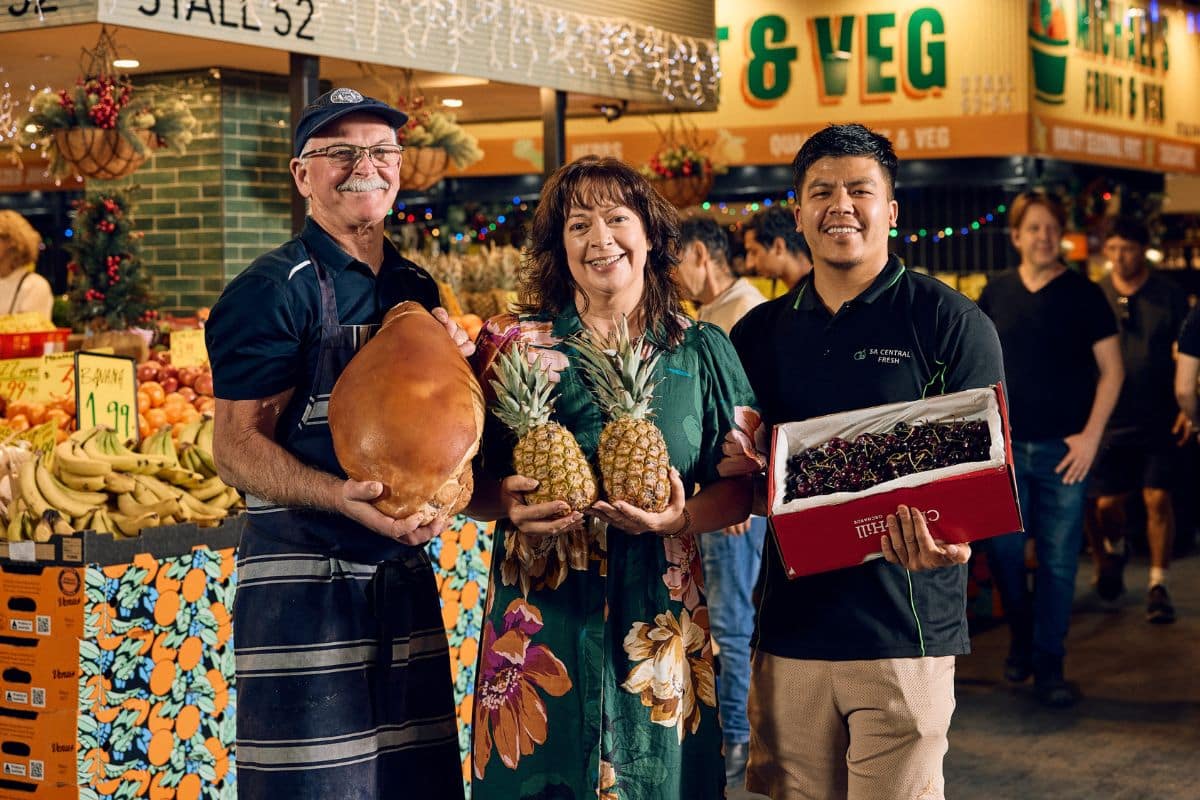 A group of three people stands before a bustling fruit and vegetable stand, presenting the assortment of fresh items offered.