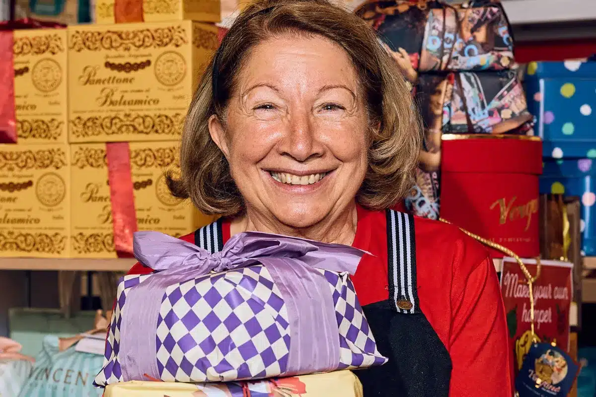 A woman joyfully holds a large box filled with colorful presents, smiling as she prepares to celebrate a special occasion.