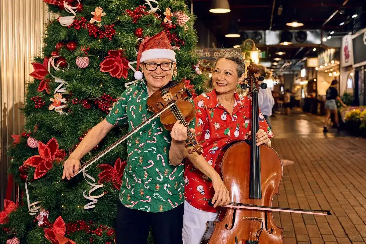  A man and woman in festive Christmas attire playing a duet on cellos, surrounded by holiday decorations.