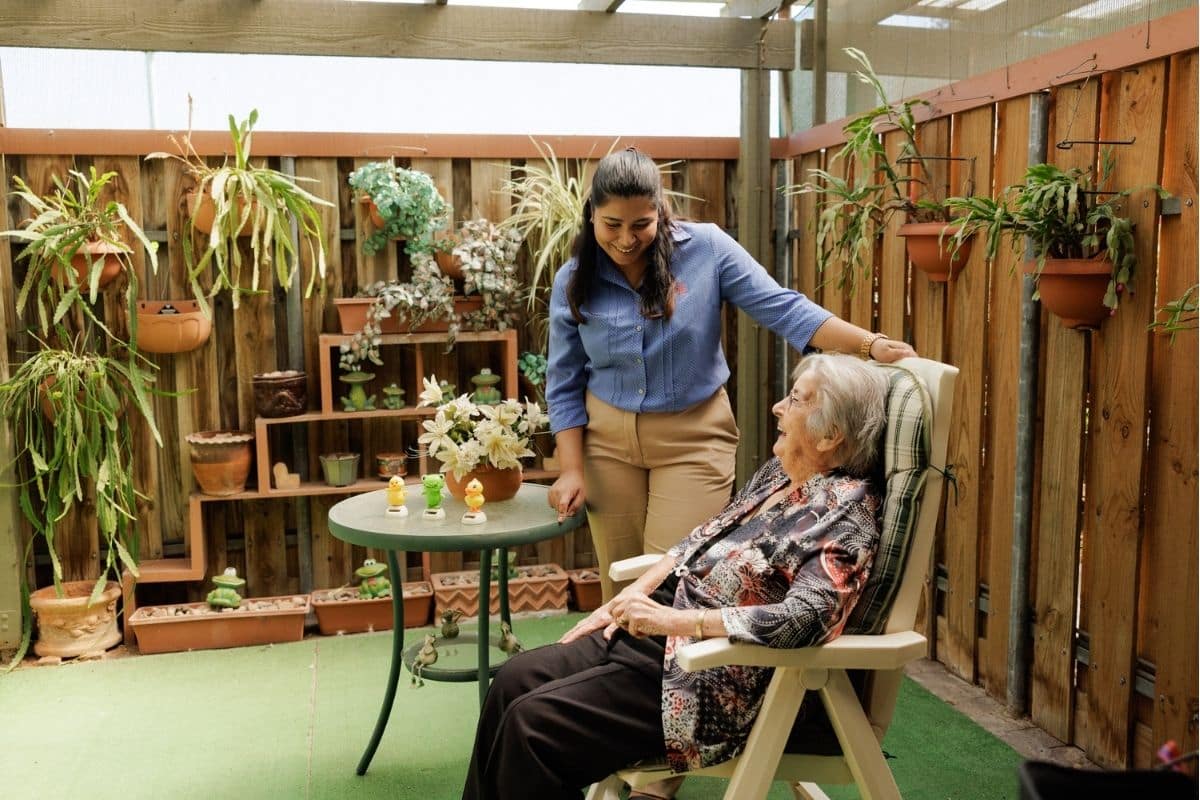 A woman assists an elderly woman seated in a chair, providing support and care in a warm garden  environment.