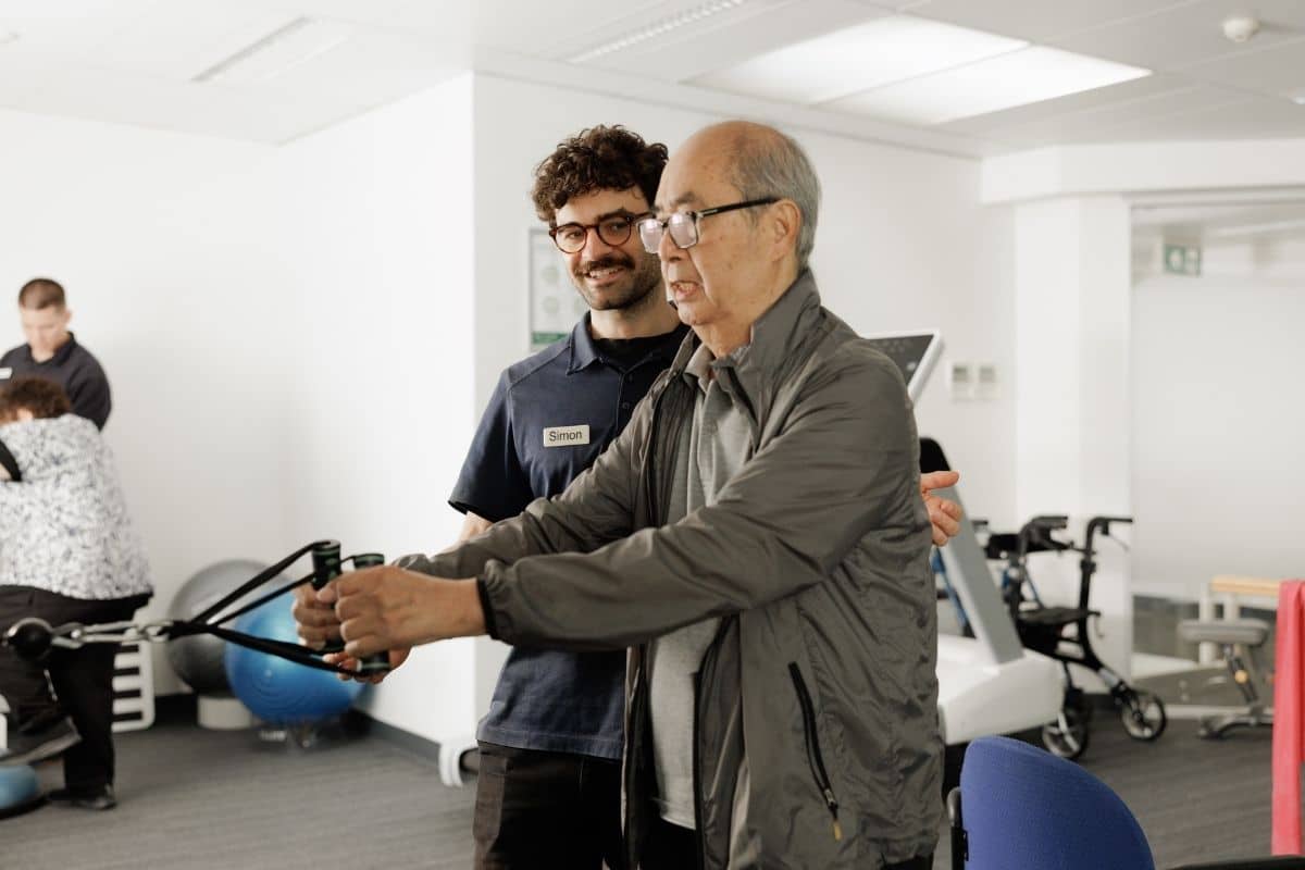 A senior man stands in a gym, gripping training equipment with both hands, focused on his workout accompanied by a trainer