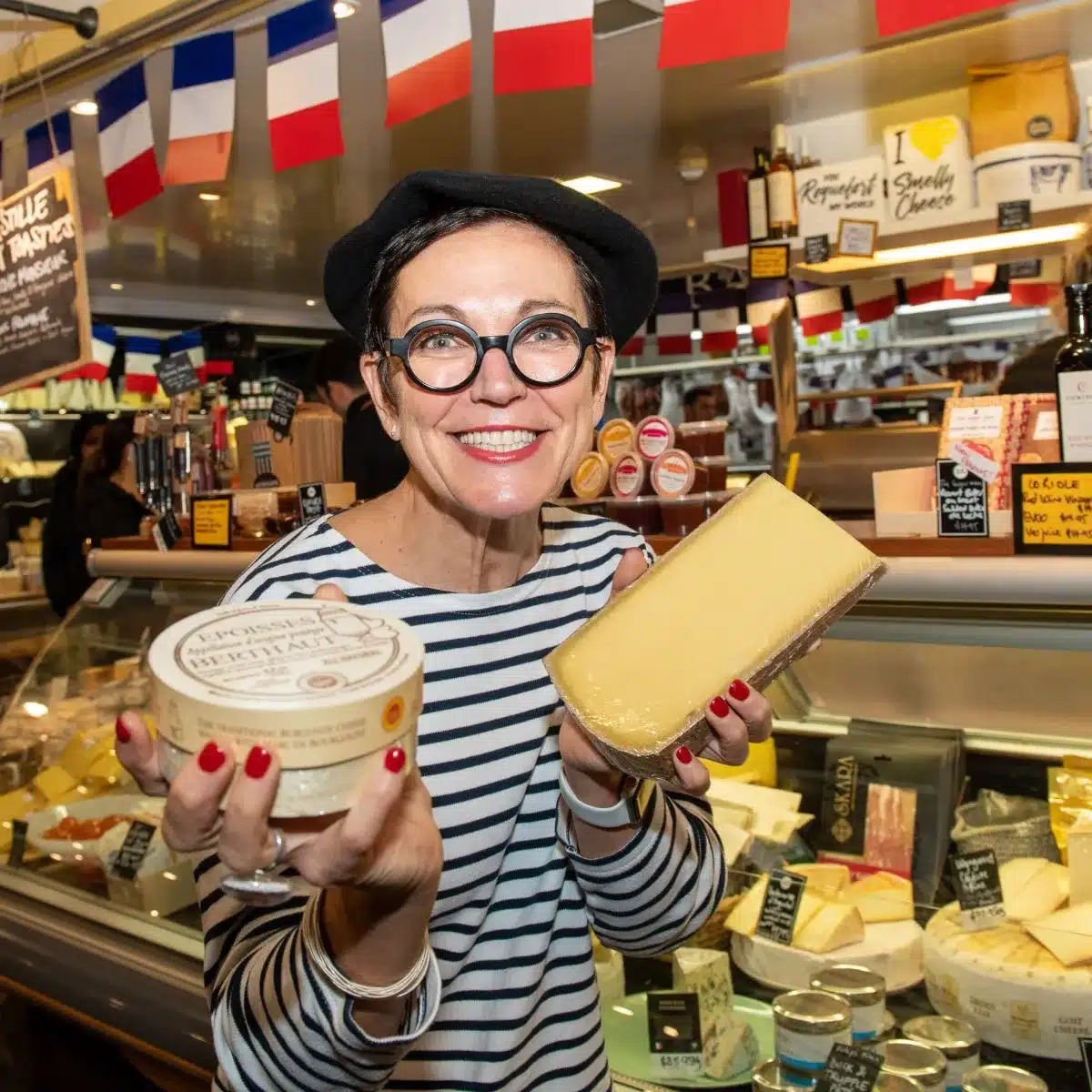 A woman dressed in a striped long sleeve t-shirt excitedly presents cheeses in front of a cheese deli counter