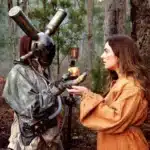 A woman in a costume offers an apple to a man in a costume, both engaged in a playful exchange advertising poster for A Concise Compendium of Wonder