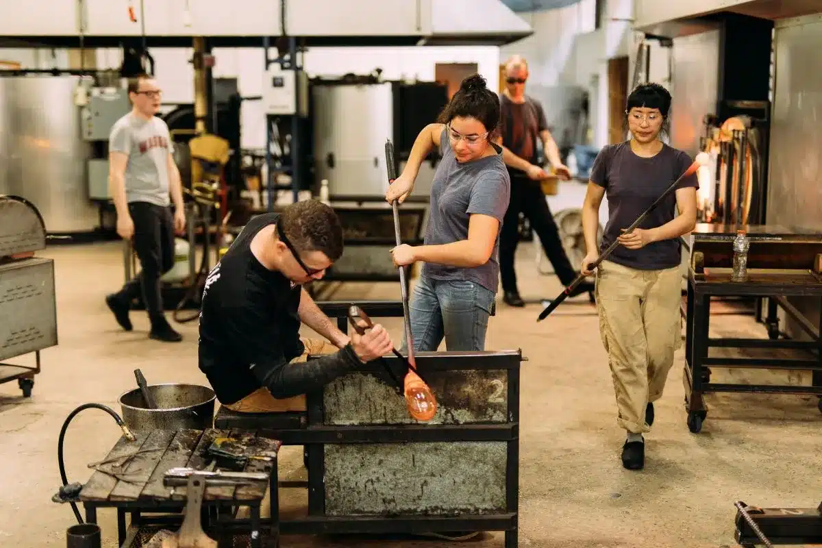Students collaboratively shaping glass in a vibrant glass blowing studio, showcasing creativity and craftsmanship.
