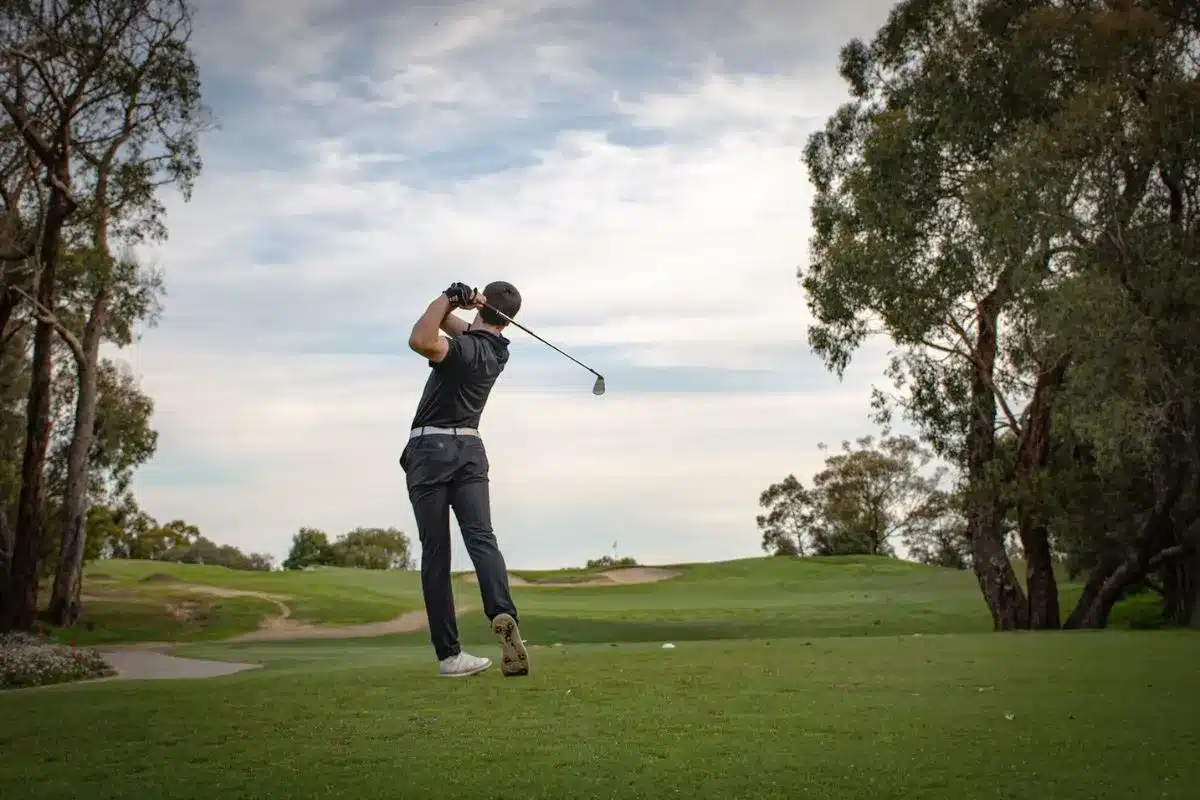 A man swings his golf club on a lush green course under clear blue skies.