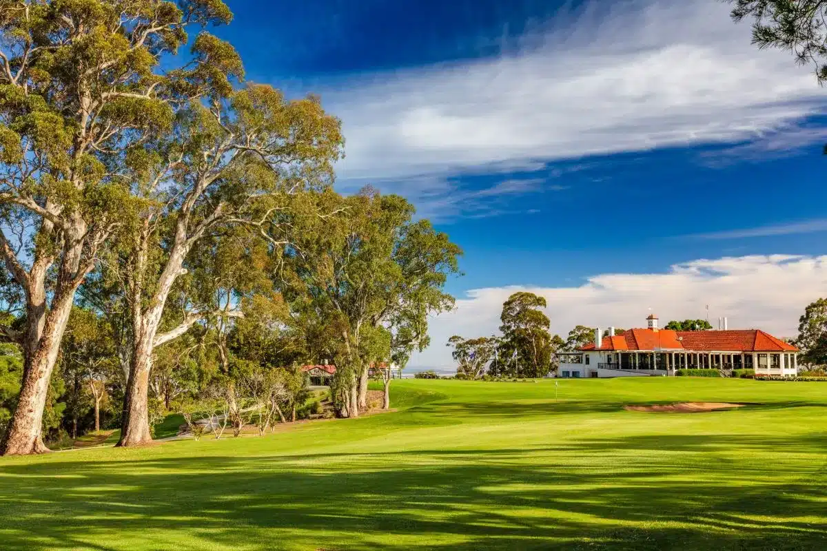 A scenic golf course featuring lush trees and a house in the background under a clear blue sky.