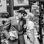 A man and woman share a kiss in front of a newspaper stand, surrounded by headlines and magazine covers.