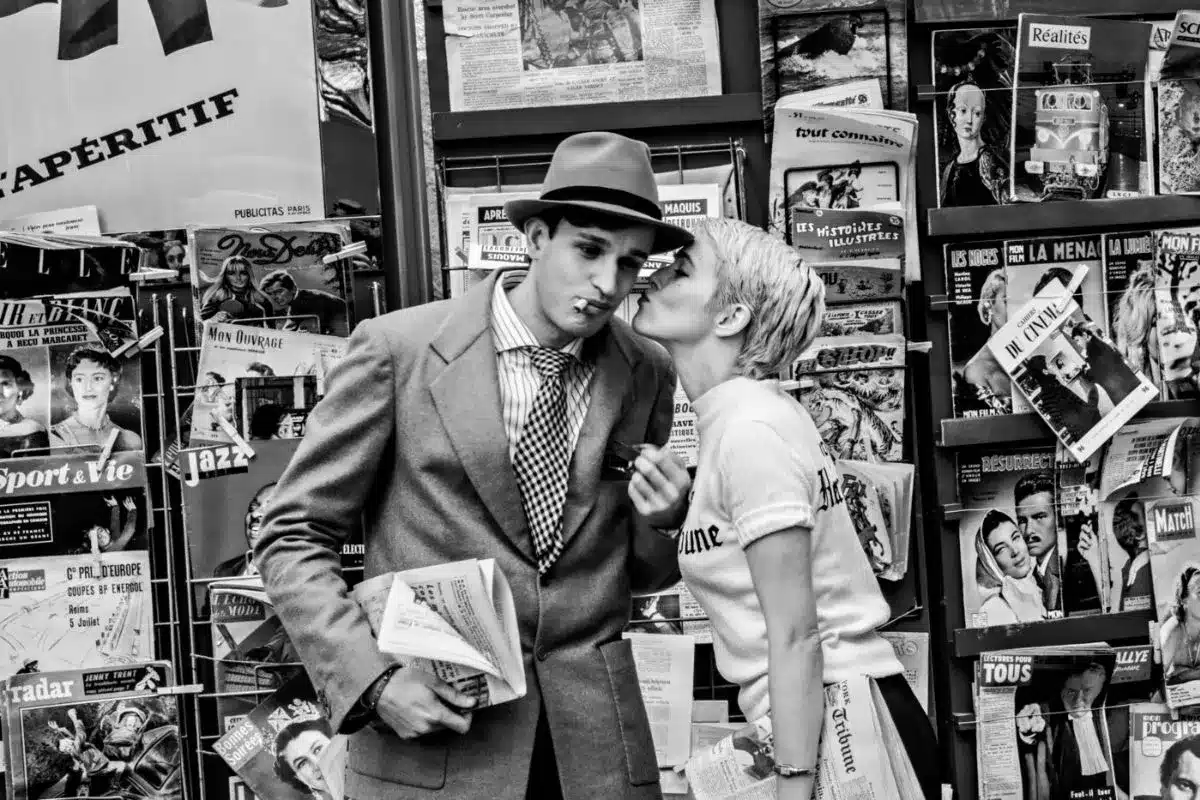 A man and woman share a kiss in front of a newspaper stand, surrounded by headlines and magazine covers.