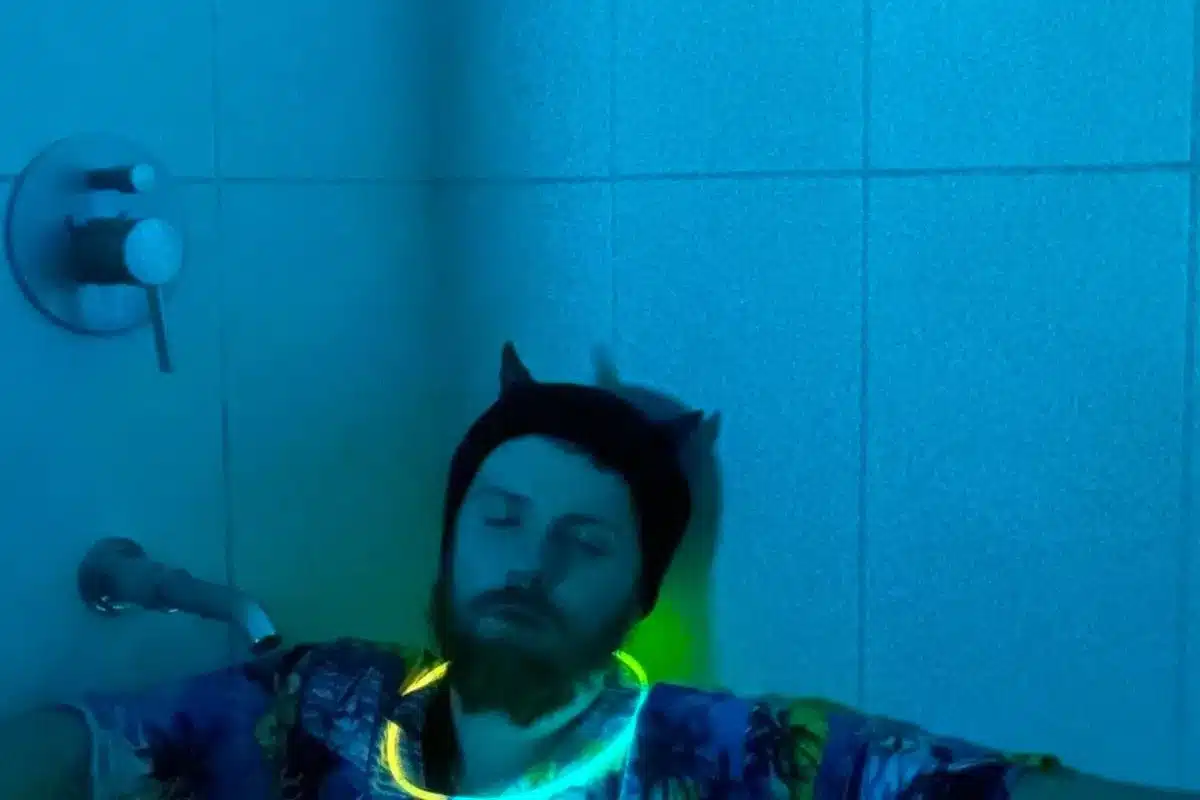 A man sits in a bath tub under blue light with glow in the dark necklace around his neck