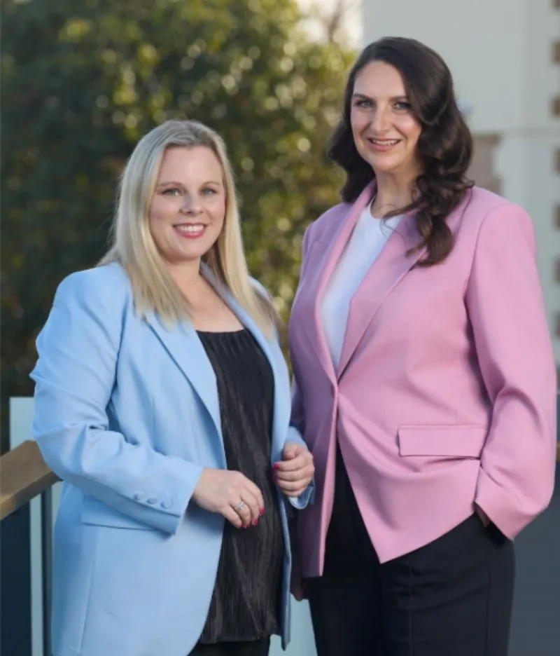 Two women in coprorate dress stand together confidently. 