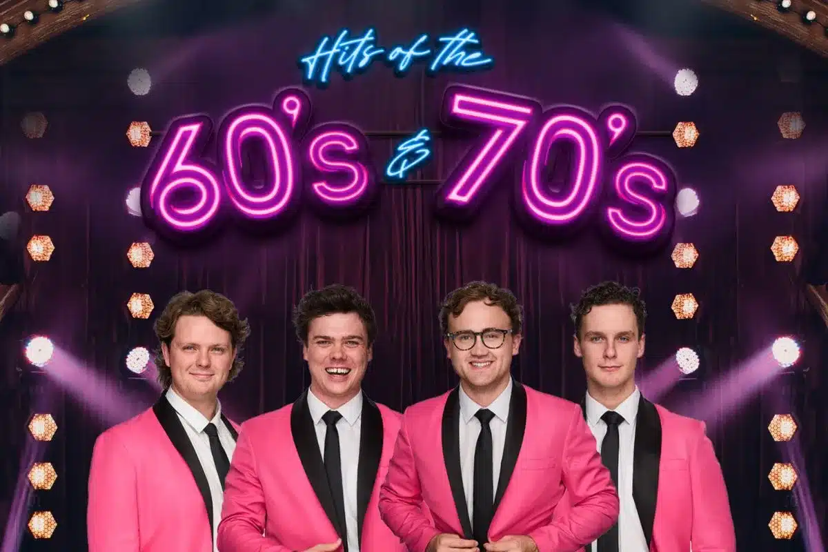 A group of four men dressed in matching hot pink coats and black ties stand under a retro neon sign 'Hits of the 60's & 70's'
