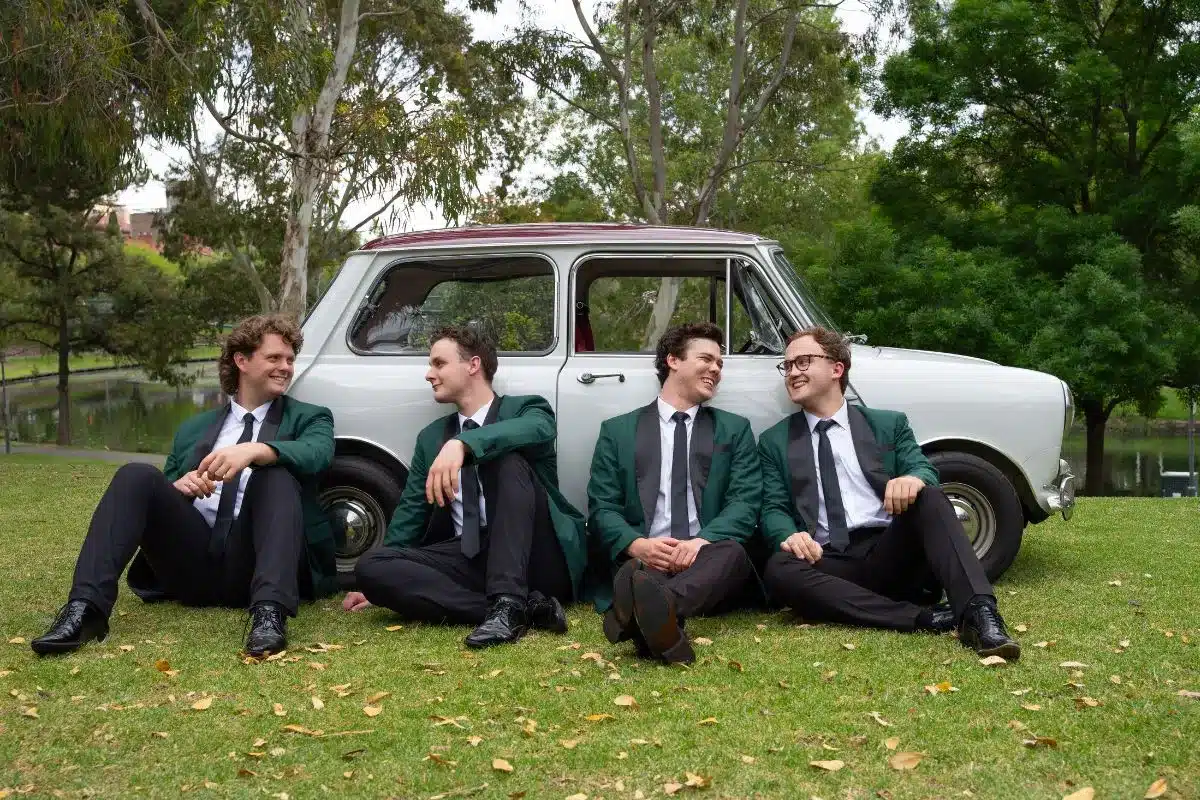 a group of 4 men dressed in matching green blazers sit by a retro car