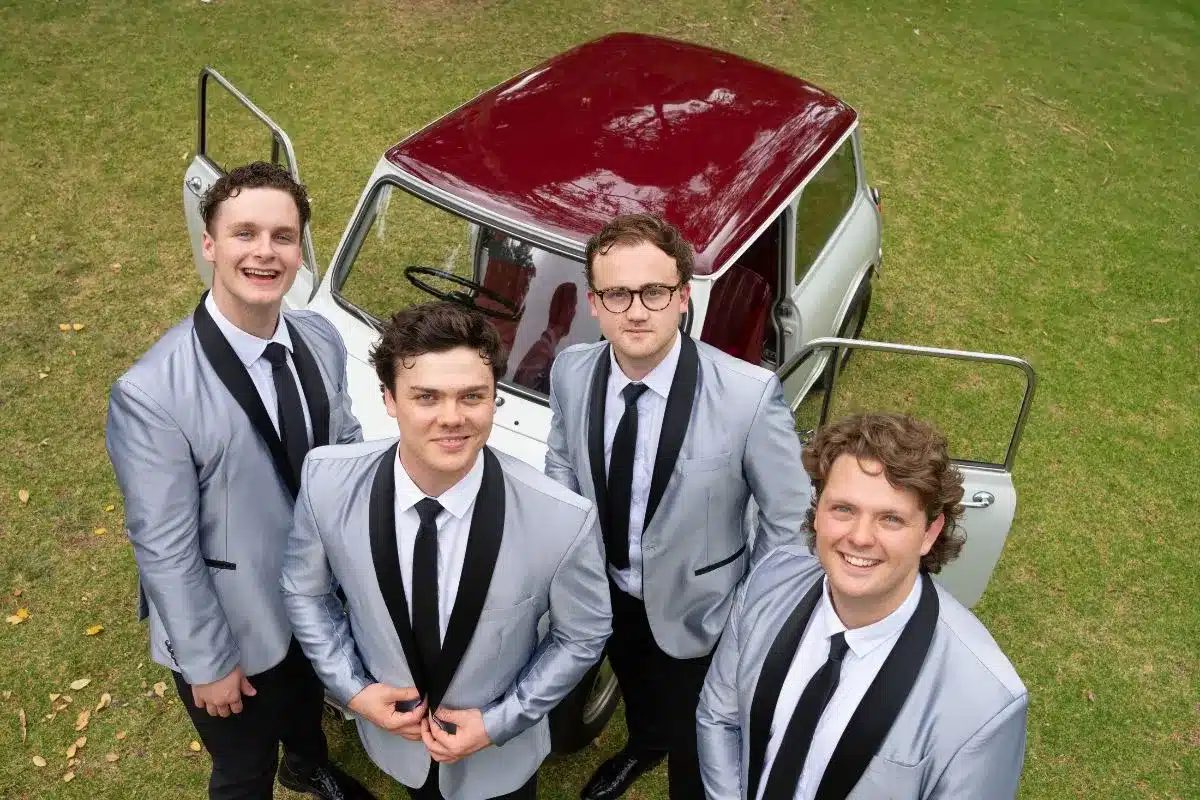 a group of 4 men dressed in matching blazers and ties stand beside a retro car looking up towards camera