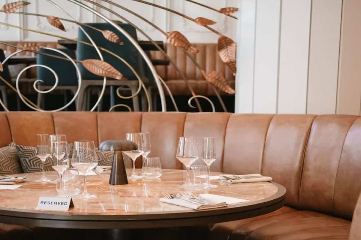 An elegantly set circular booth with tan leather banquette seating, marble-top table, crystal wine glasses and a reserved sign, with decorative copper leaf metalwork in the background