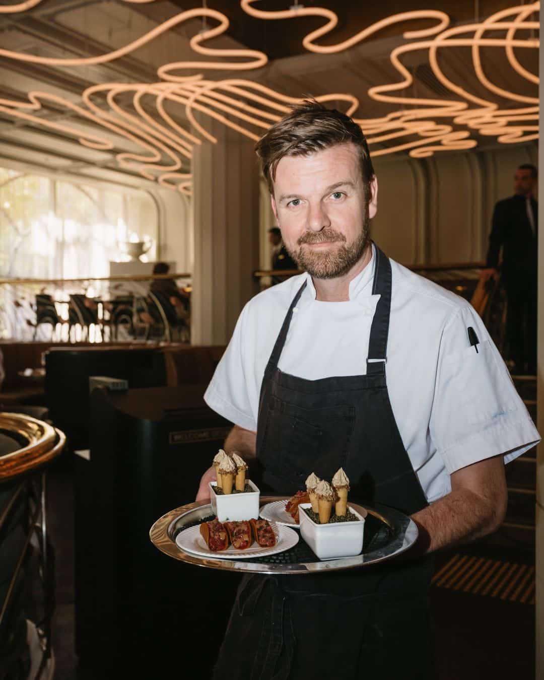 A chef in a white jacket and black apron holds a silver tray of elegant canapés, standing beneath swirling copper neon lights in a stylish restaurant