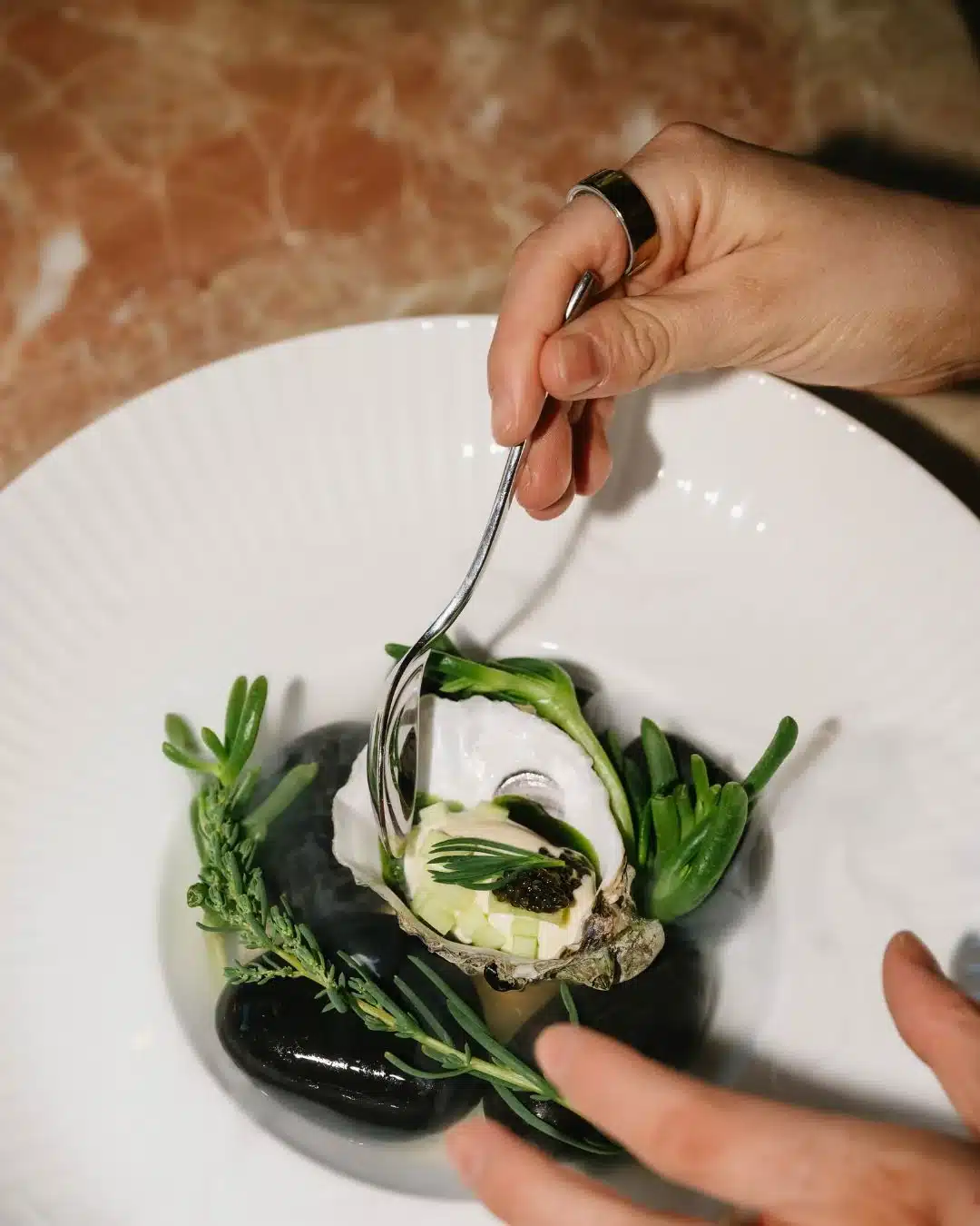 A hand using a silver spoon to dress a freshly shucked oyster with caviar, served on black stones and surrounded by fresh coastal herbs and sea succulents on a white ridged plate