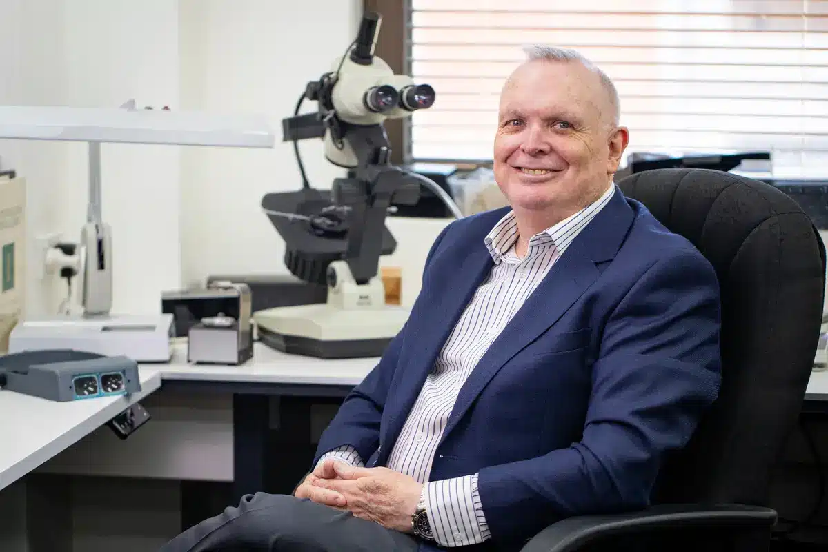 Gerard McCabe seated in his workshop wearing a navy blazer, smiling warmly at the camera, with a gemological microscope and jewellery tools visible on the workbench behind him.