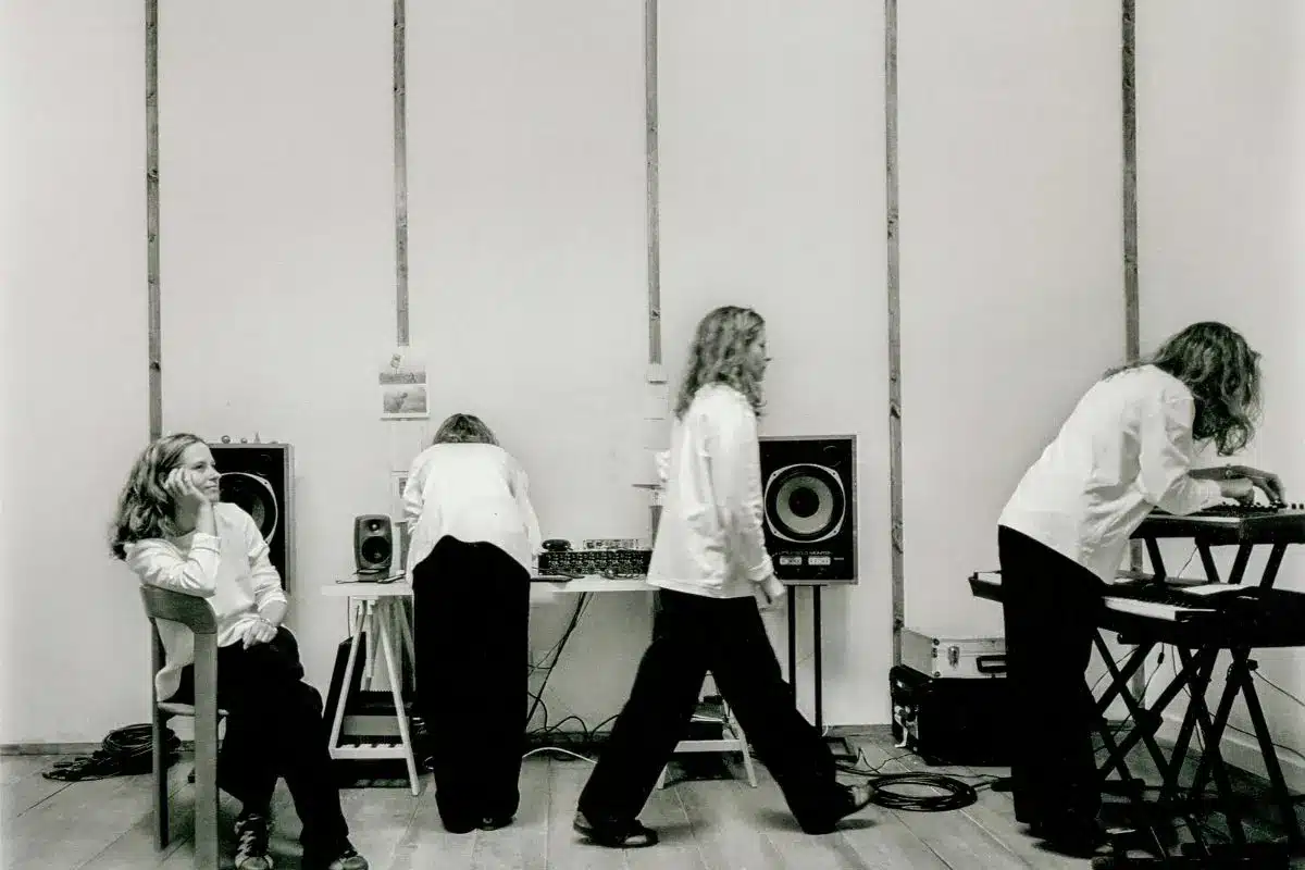 Black and white photograph of four figures dressed in white shirts and black trousers at various positions around audio equipment and speakers in a sparse, minimalist room — an artistic performance or rehearsal setting.
