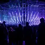 Silhouettes of a crowd standing before a large immersive light installation featuring cascading vertical LED tubes glowing in vivid purple and blue, part of the Illuminate Adelaide City Lights program.