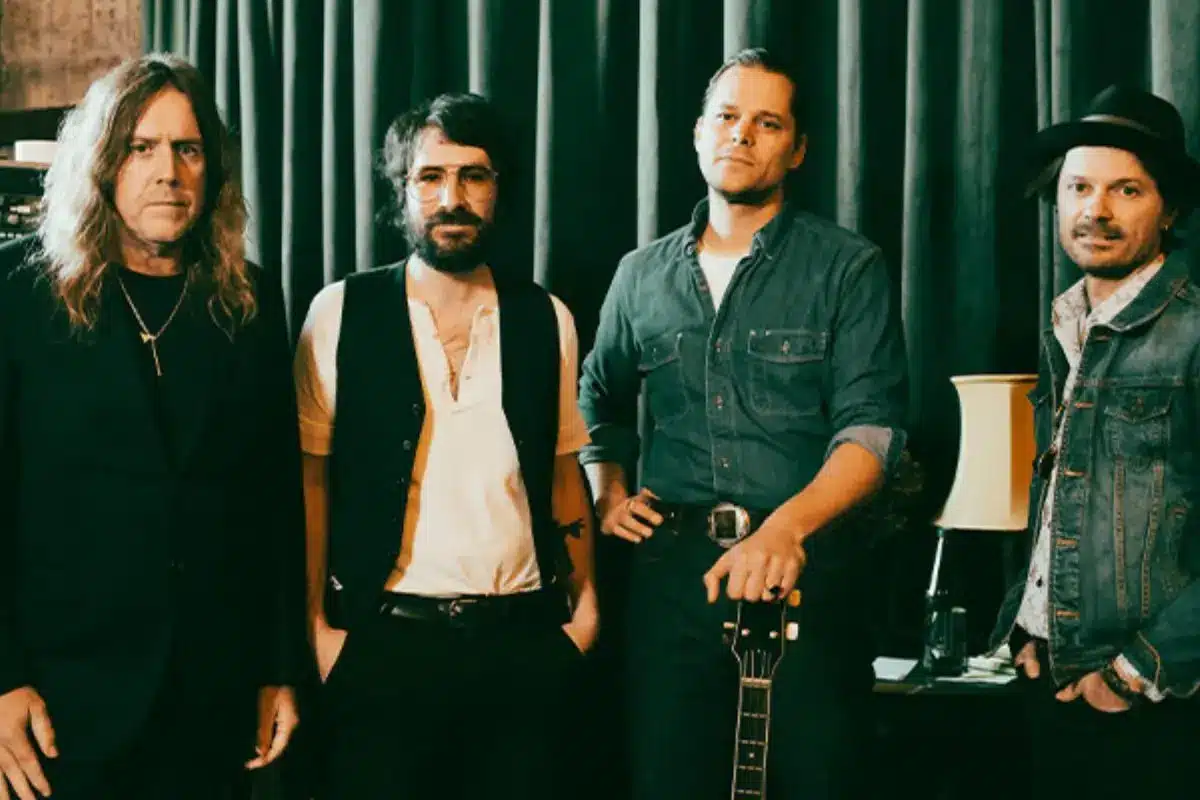 Four members of rock band ARC standing in front of a dark teal curtain, one holding a guitar by the neck. The group is styled in a moody, vintage rock aesthetic.