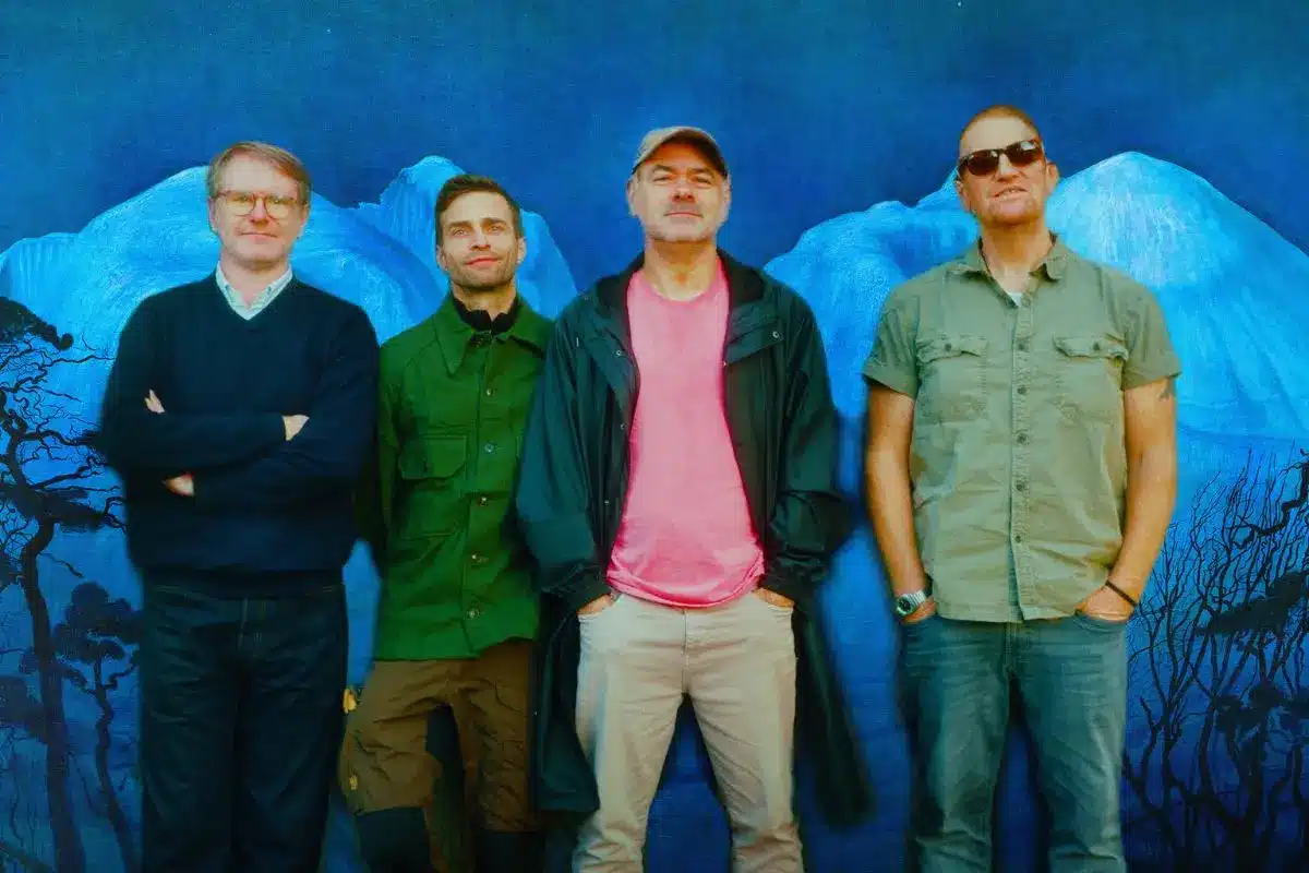 Four members of The Beta Band standing casually in front of a large blue painted mural depicting mountains and bare trees.