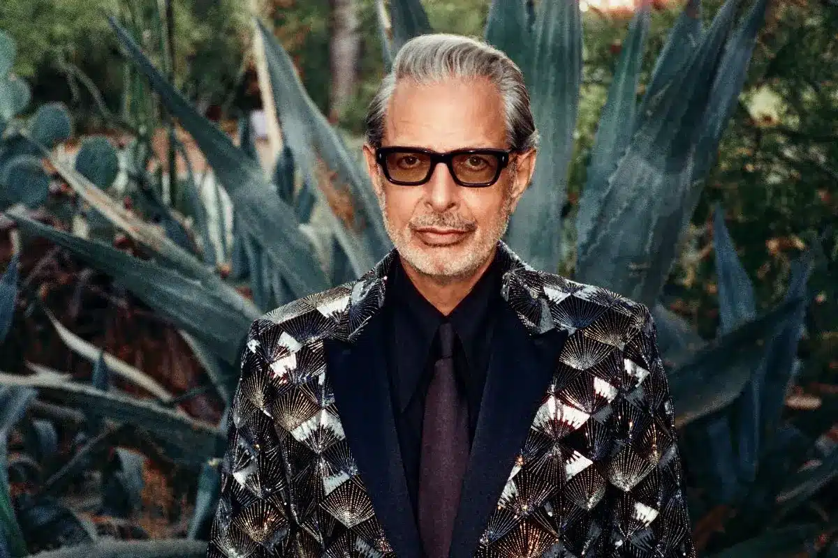 Portrait of Jeff Goldblum wearing a patterned sequined blazer and dark sunglasses, posed in front of large agave plants.