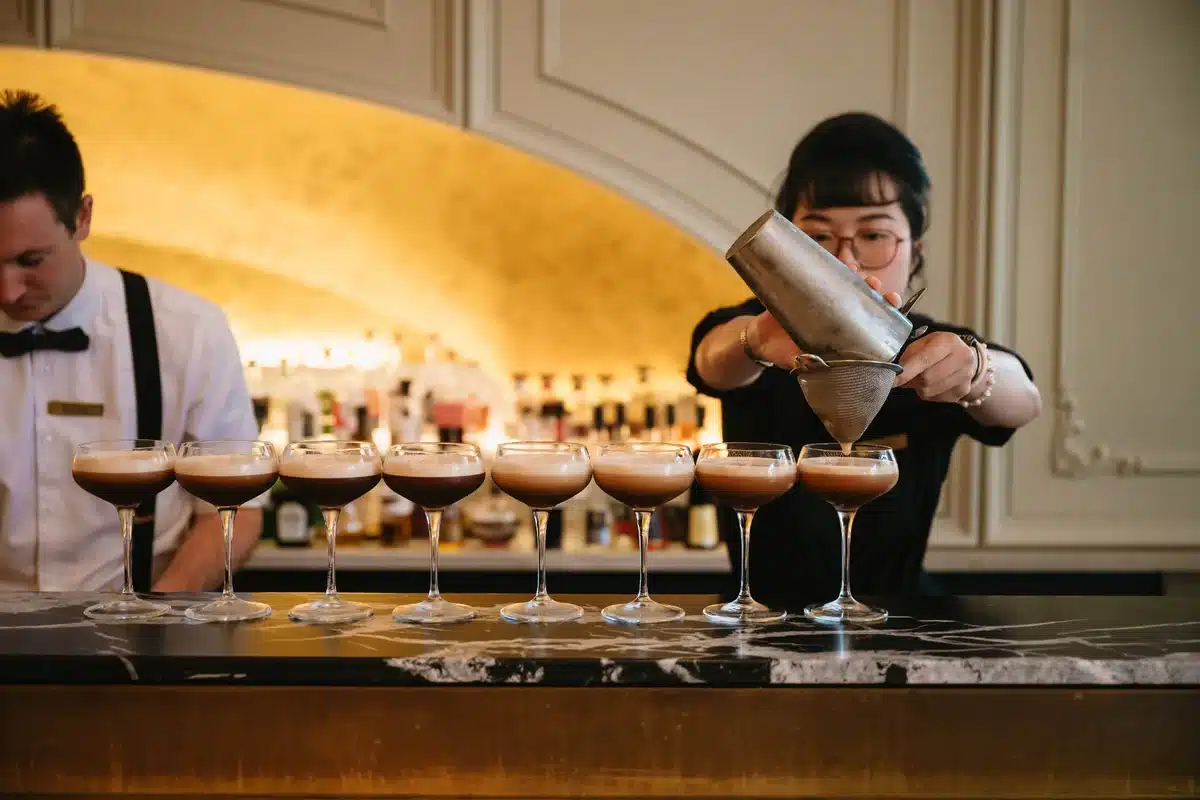 A bartender pouring a row of cocktails through a strainer at a marble bar, with a second bartender working in the background against a warmly lit gold arch