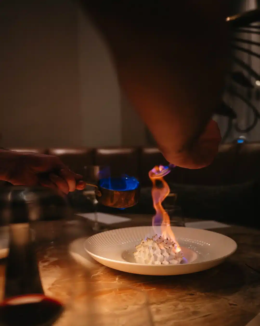 A meringue dessert being dramatically flambéed tableside, with blue and orange flames rising from a copper pan over a white bowl on a marble surface