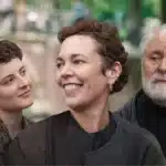 Promotional image for the film Jimpa featuring three characters against a blurred green outdoor backdrop — a young curly-haired woman on the left, a smiling woman with short auburn hair in the centre, and an older bearded man on the right.