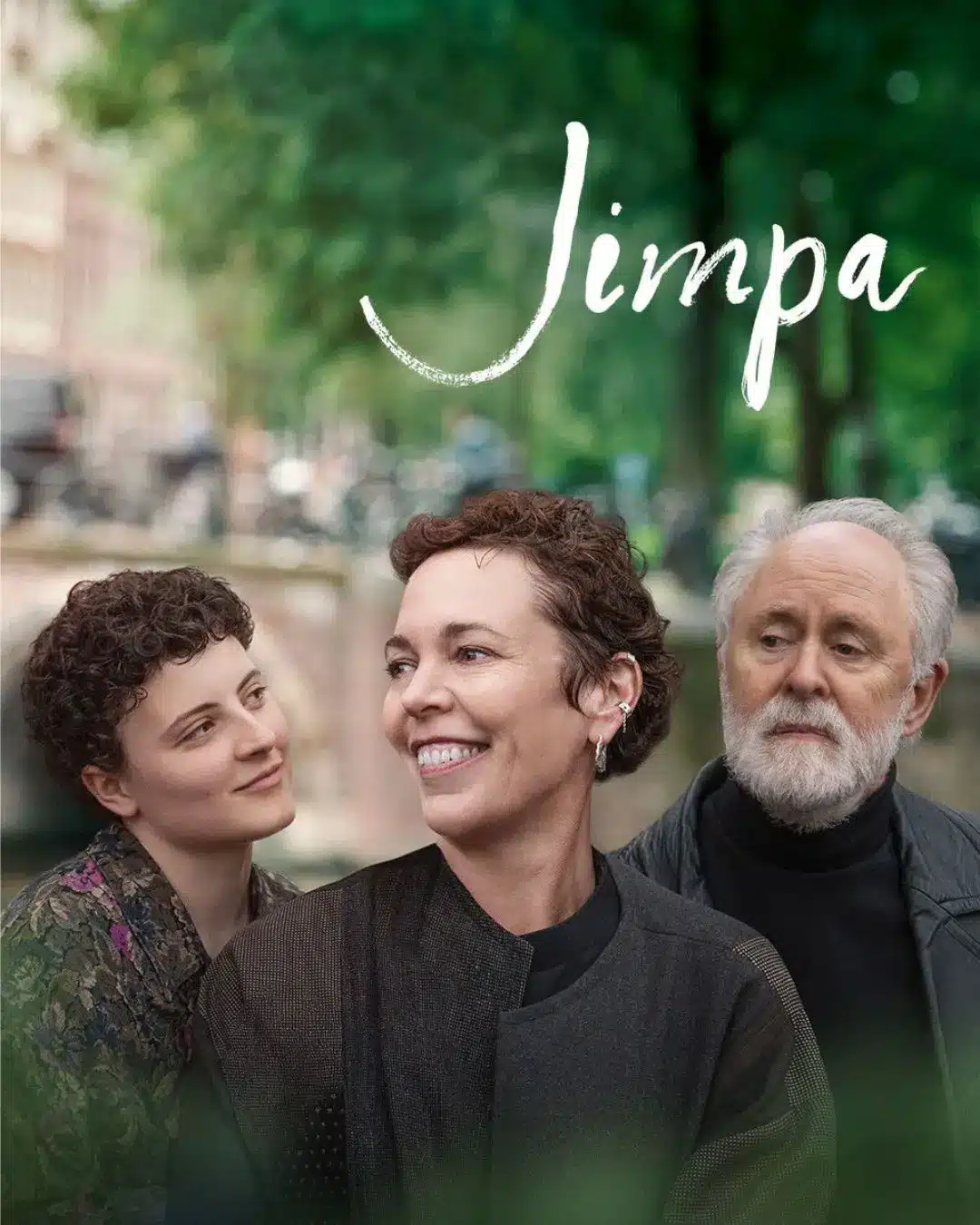Promotional image for the film Jimpa featuring three characters against a blurred green outdoor backdrop — a young curly-haired woman on the left, a smiling woman with short auburn hair in the centre, and an older bearded man on the right.