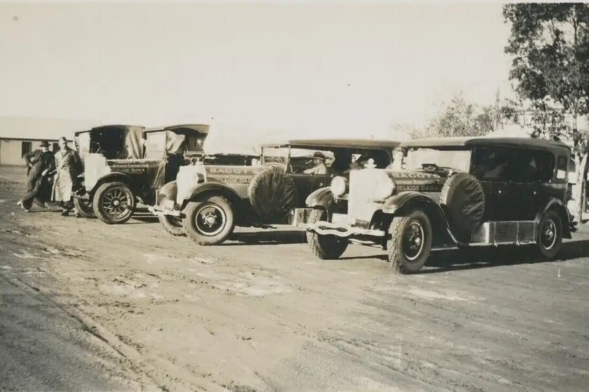 A sepia-toned archival photograph of three Bagot's Adelaide–Darwin touring cars lined up on a dirt road, with passengers and bystanders gathered around them.